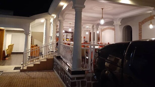 A covered outdoor area with a staircase leading to an upper level, white pillars, and hanging lights. - Cari Hotel (Kigali)