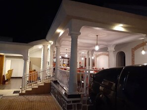 A covered outdoor area with a staircase leading to an upper level, white pillars, and hanging lights. - Cari Hotel (Kigali)