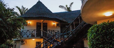 A building with a thatched roof, a staircase leading to a balcony, and a well-lit entrance.