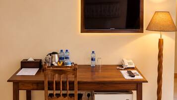 A hotel room with a wooden desk, a chair, a television mounted on the wall, a desk lamp, a mini-fridge, and an air conditioning unit.