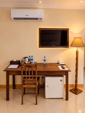 Une chambre d’hôtel équipée d’un bureau en bois, d’une chaise, d’un téléviseur fixé au mur, d’une lampe de bureau, d’un mini-réfrigérateur et d’un climatiseur.