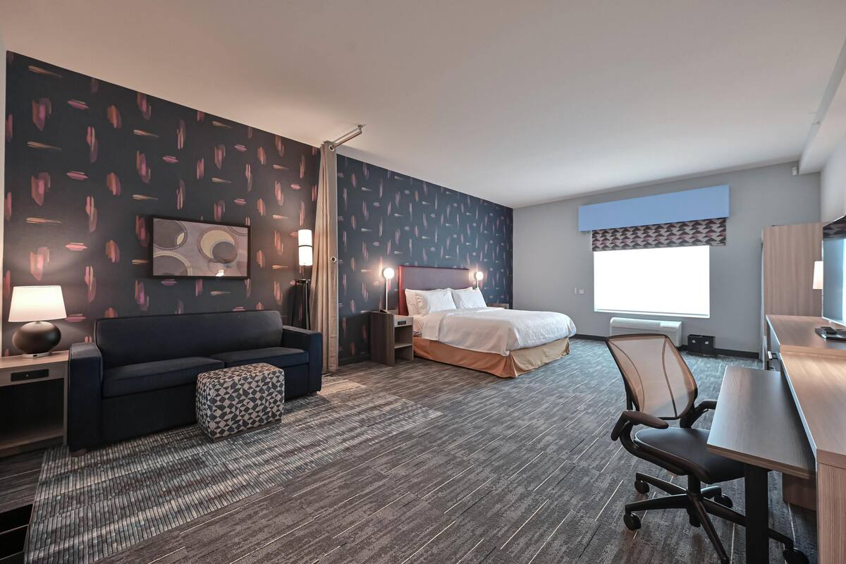 Quarto de hotel com cama, escrivaninha, cadeira, sofá, luminária e janela.