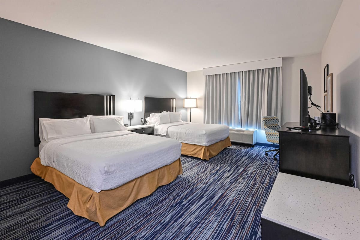 Quarto de hotel com duas camas, uma escrivaninha, uma cadeira, televisão e janela com cortinas.