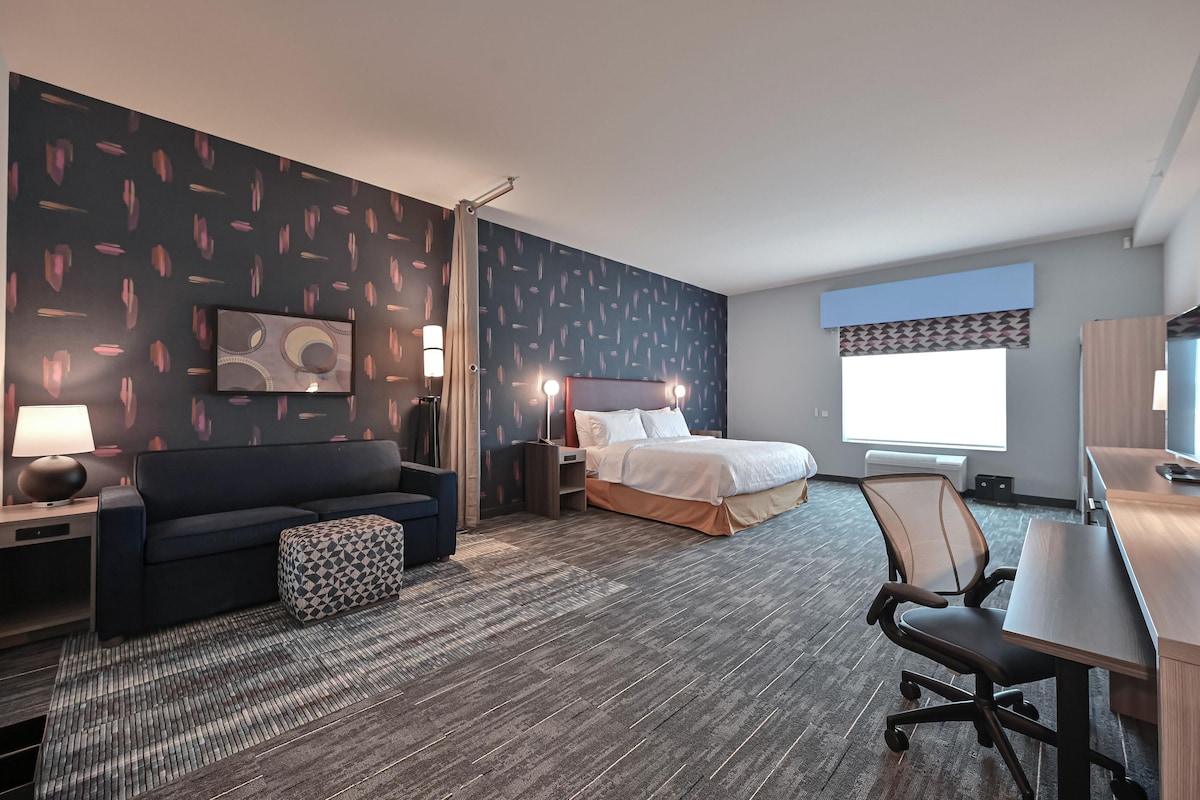 A hotel room with a bed, a desk, a chair, a sofa, a lamp, and a window.