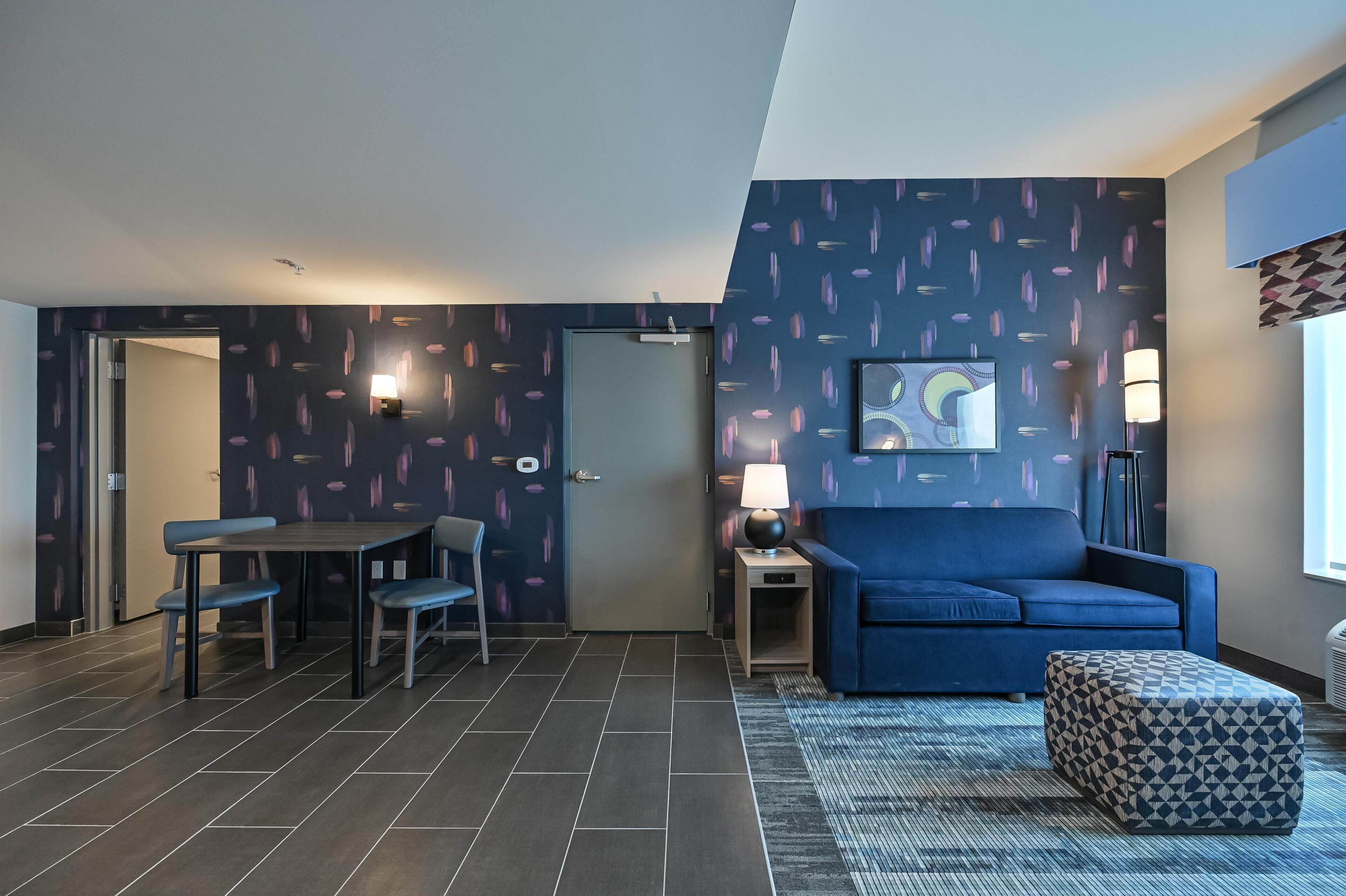 A modern room with a dark patterned wall, a blue sofa, a small table with chairs, a lamp, and a rug.