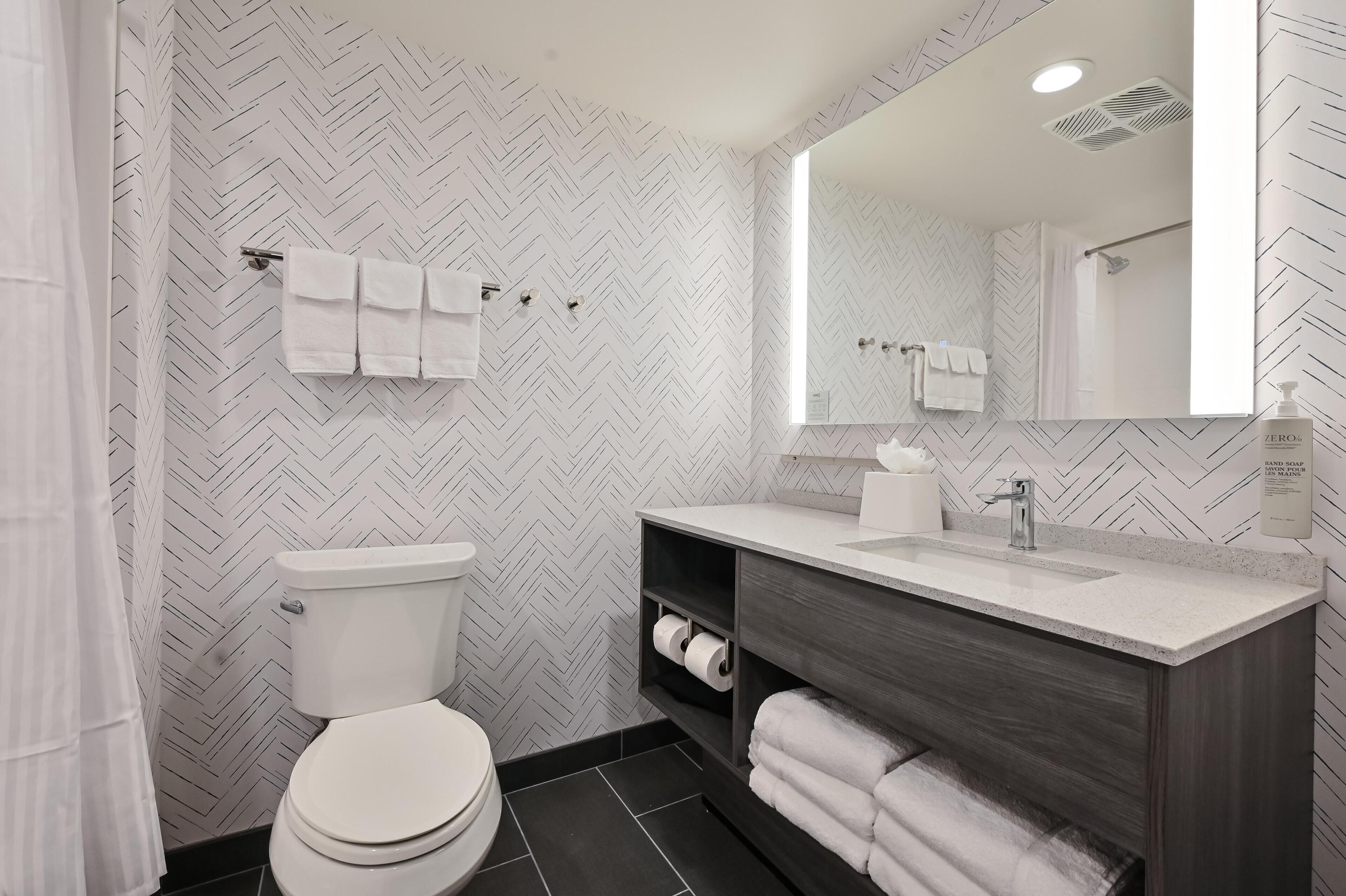 Une salle de bain moderne avec un mur orné d’un motif en chevrons, des toilettes, un lavabo avec un miroir et un porte-serviettes.