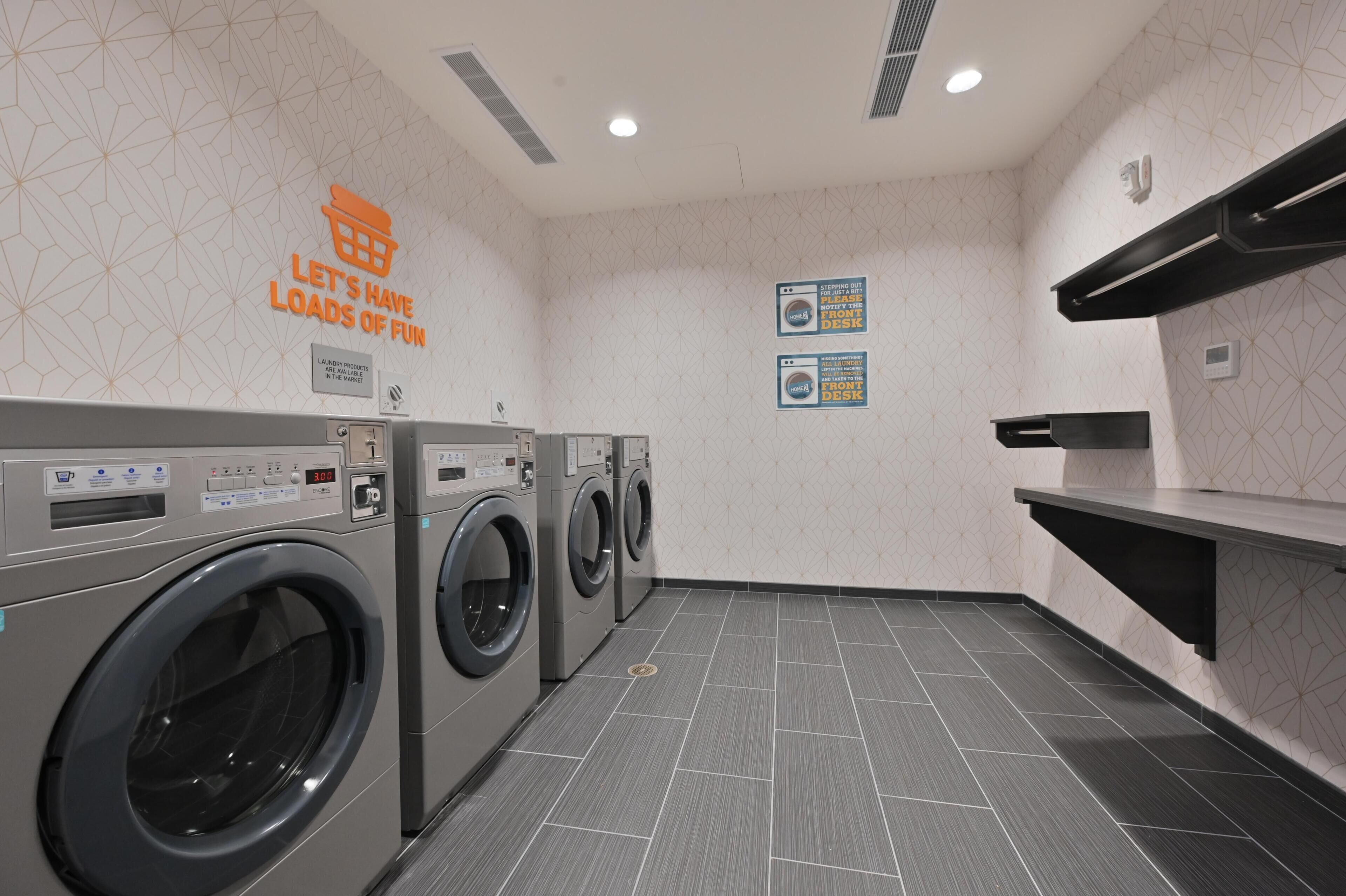 Une buanderie équipée de plusieurs machines à laver, d’un étendoir mural et d’un panneau indiquant la disponibilité des produits de lessive.
