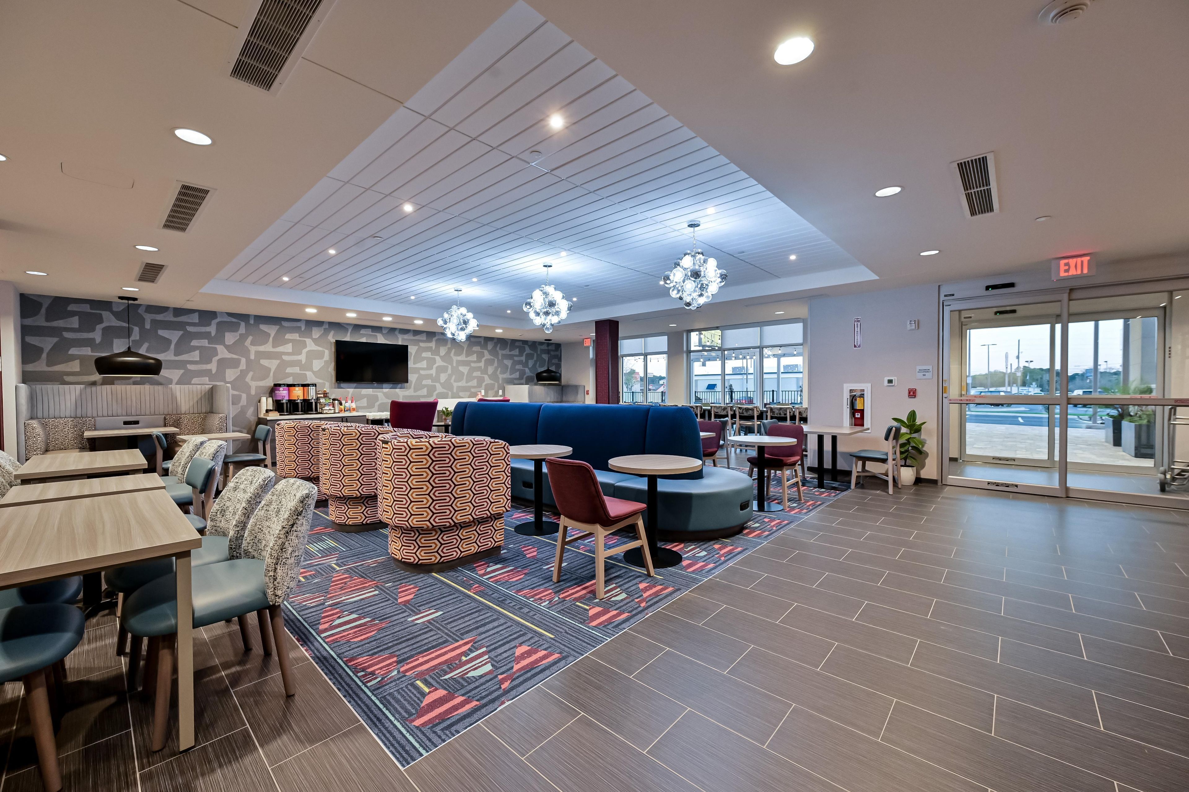 A modern hotel lobby with seating areas, a flat-screen TV, and a view through the glass doors.
