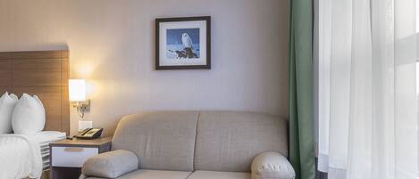 Habitación de hotel con cama, sofá, mesita de noche con lámpara, cuadro en la pared y ventana con cortinas translúcidas.