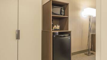 A compact kitchenette with a microwave and refrigerator, a small shelf with snacks, and a floor lamp.