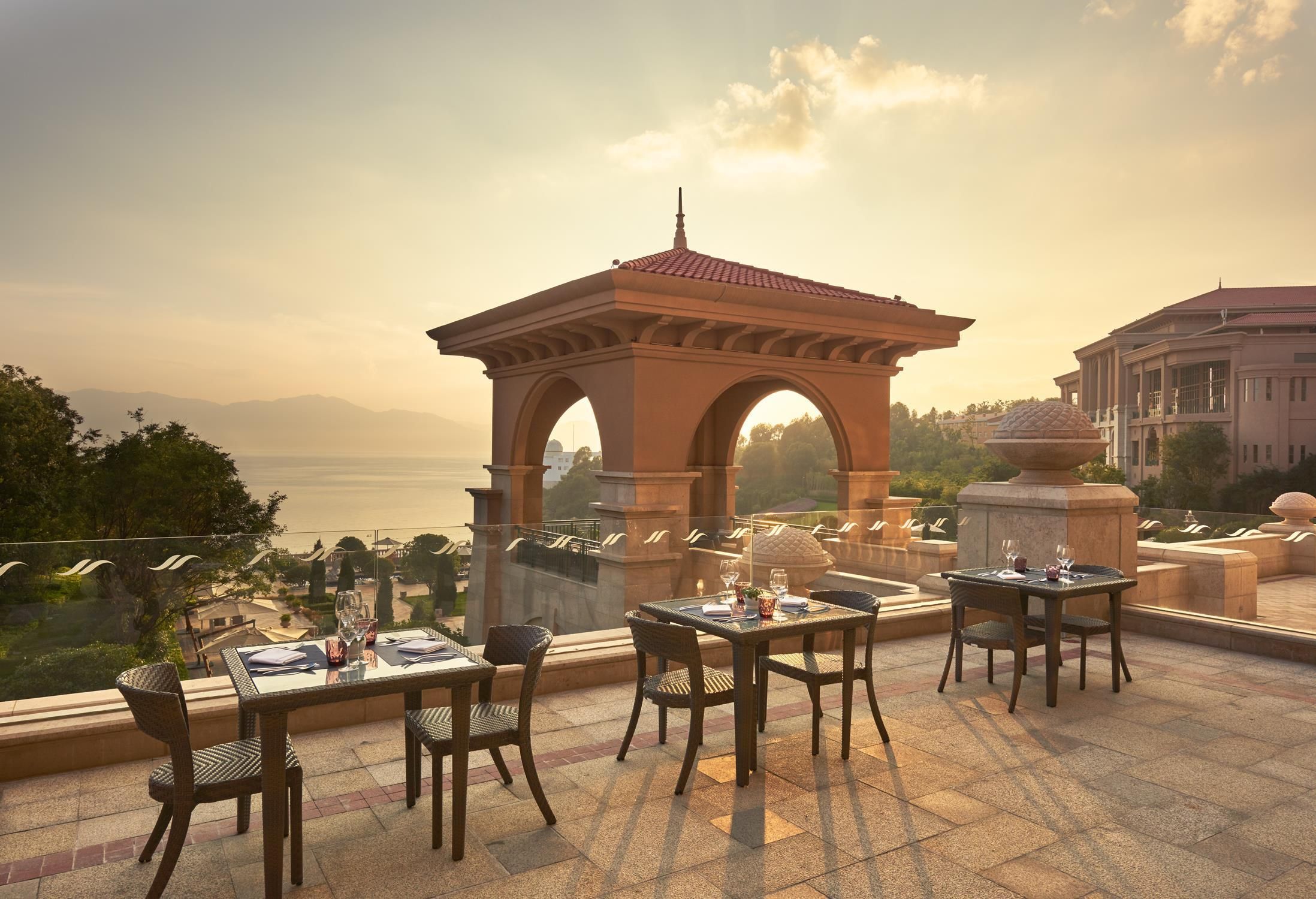 A terrace with tables and chairs, overlooking a body of water and a distant mountain range.