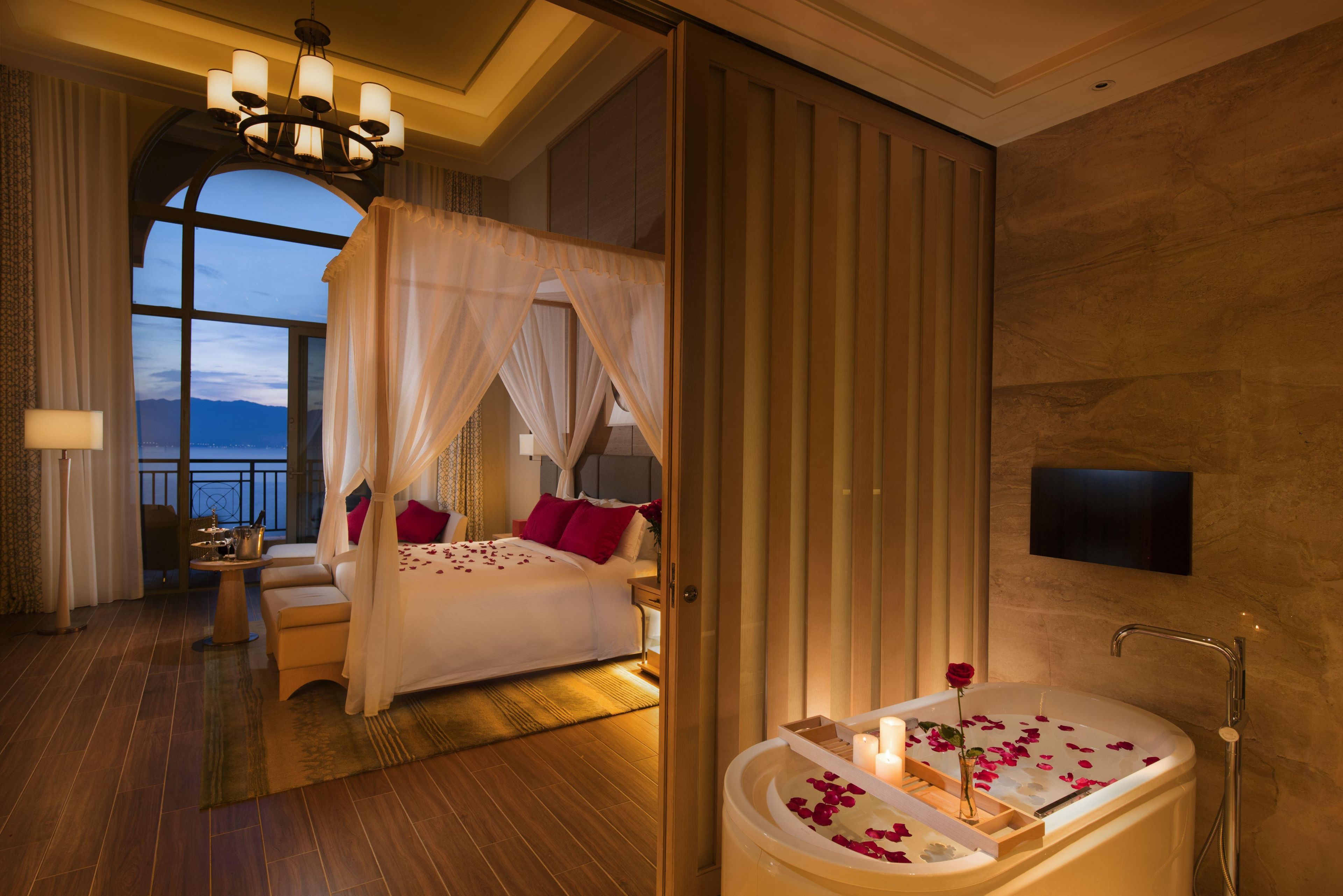 A luxurious bedroom with a four-poster bed, a large window, a sitting area with a view, and a bathtub with rose petals.