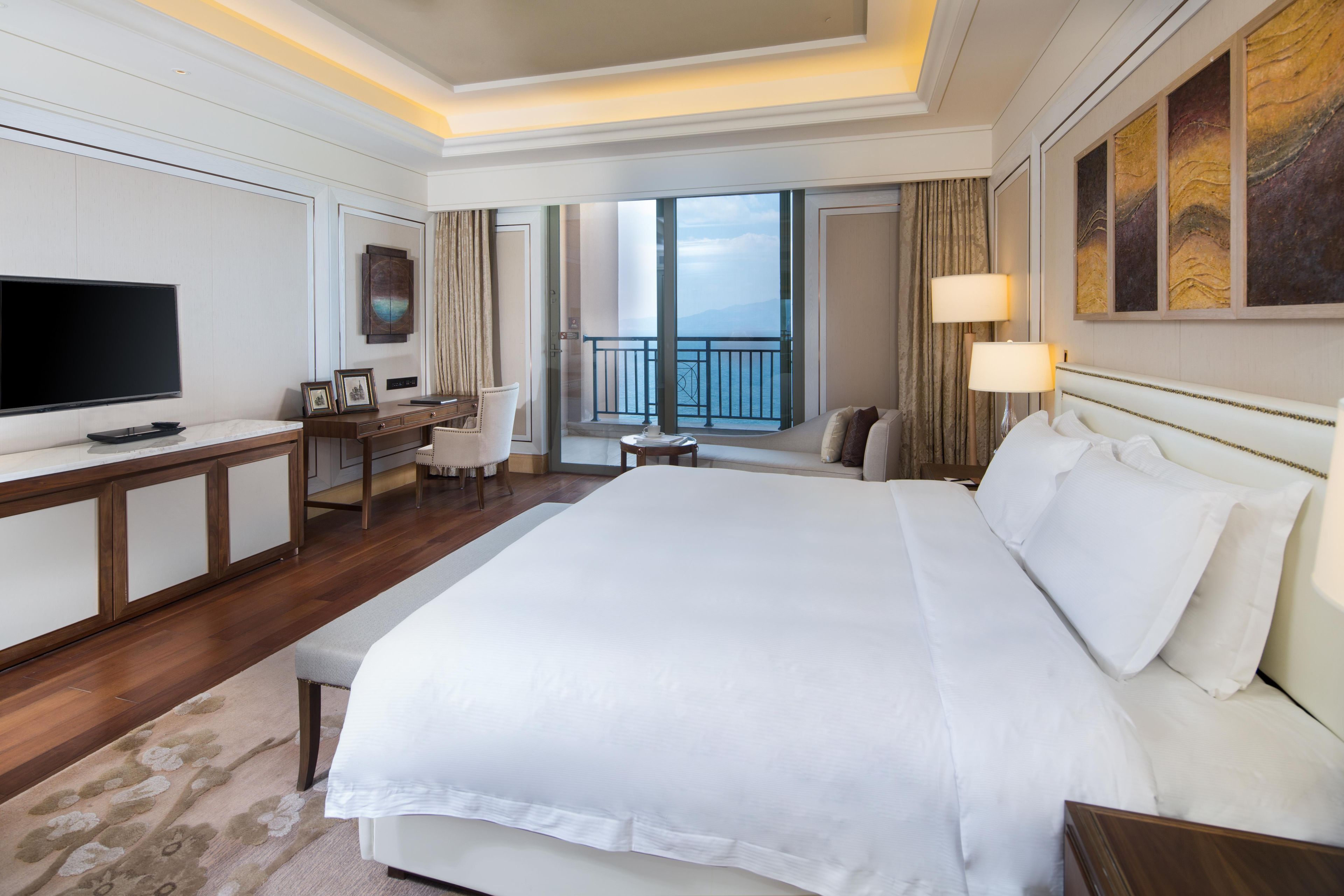 A hotel room with a large bed, a desk with a flat-screen TV, a balcony with a view, and a large painting on the wall.