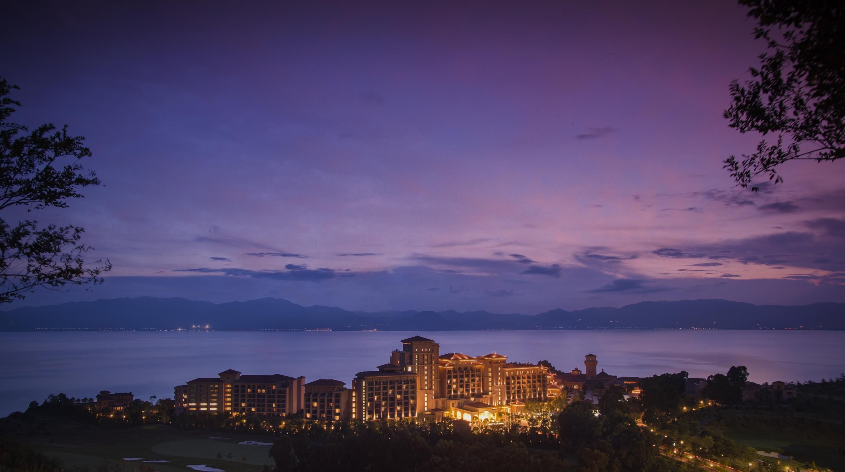 Un complejo hotelero con varias edificaciones, una masa de agua al fondo y un paisaje montañoso.