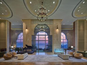 A spacious lobby with a grand piano, comfortable seating, a glass display case, and a large arched window with a sunset view.