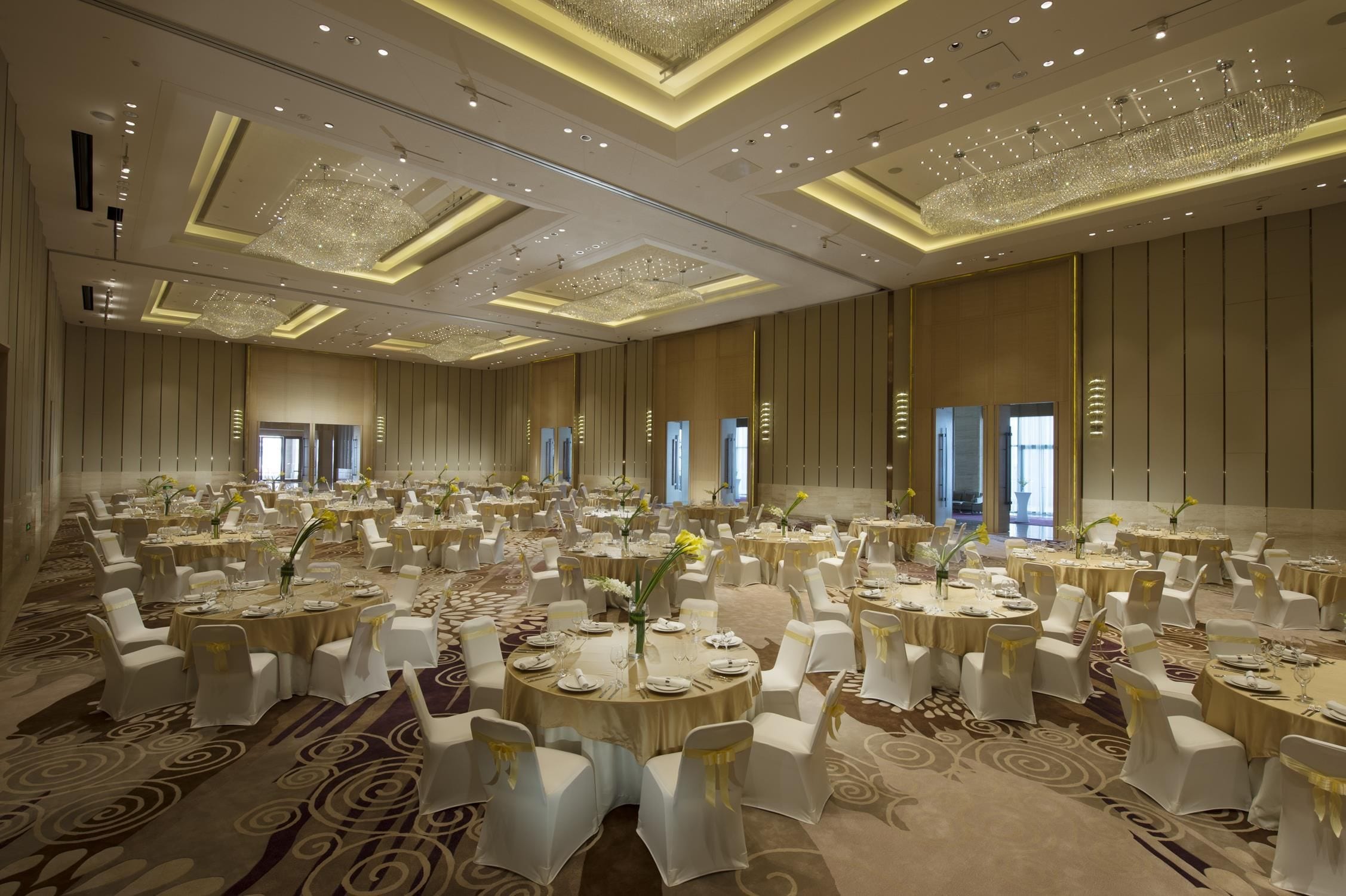 A large banquet hall with round tables, white chairs, and chandeliers.