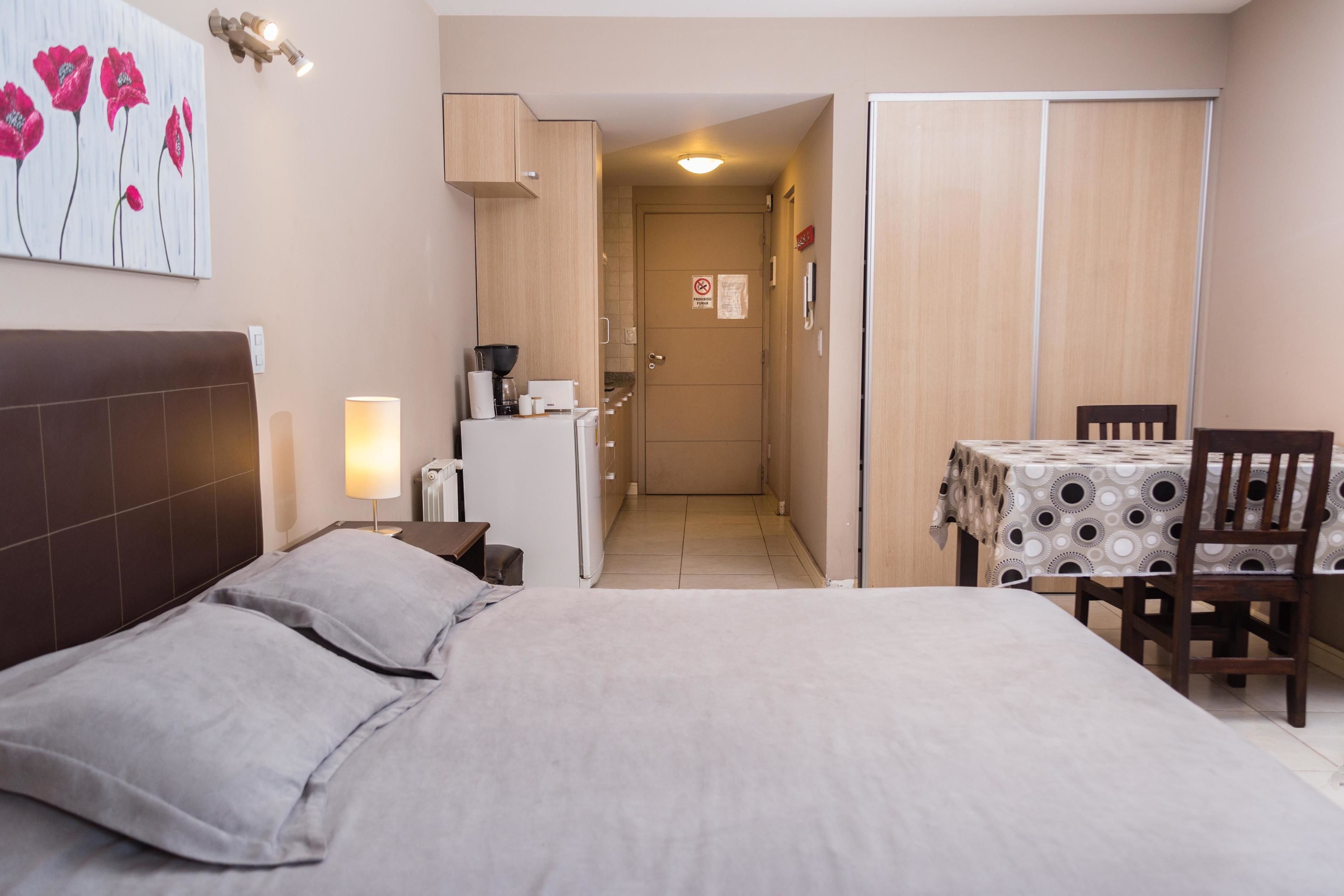 Una habitación de hotel con cama, una mesa con sillas, un kitchenette y una puerta que da al pasillo.