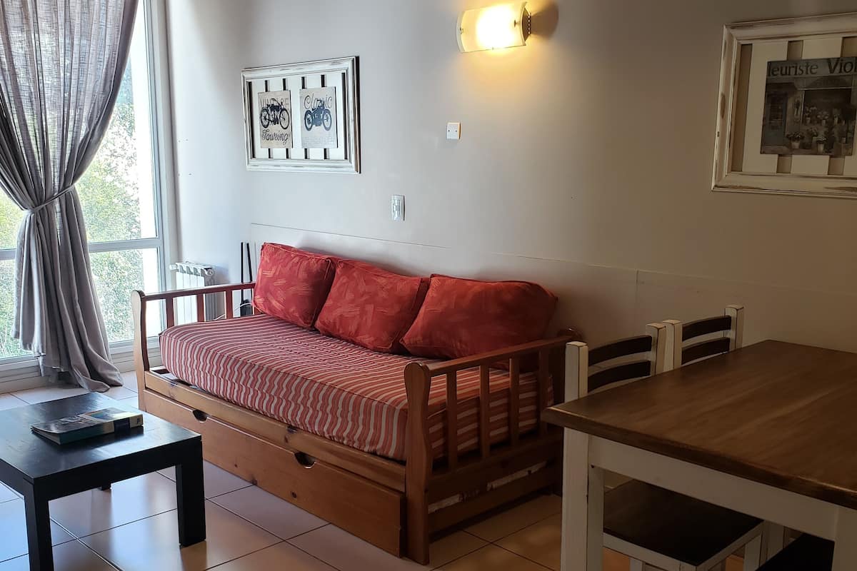 A cozy room with a wooden sofa, a small table, a dining table, and a window with curtains.