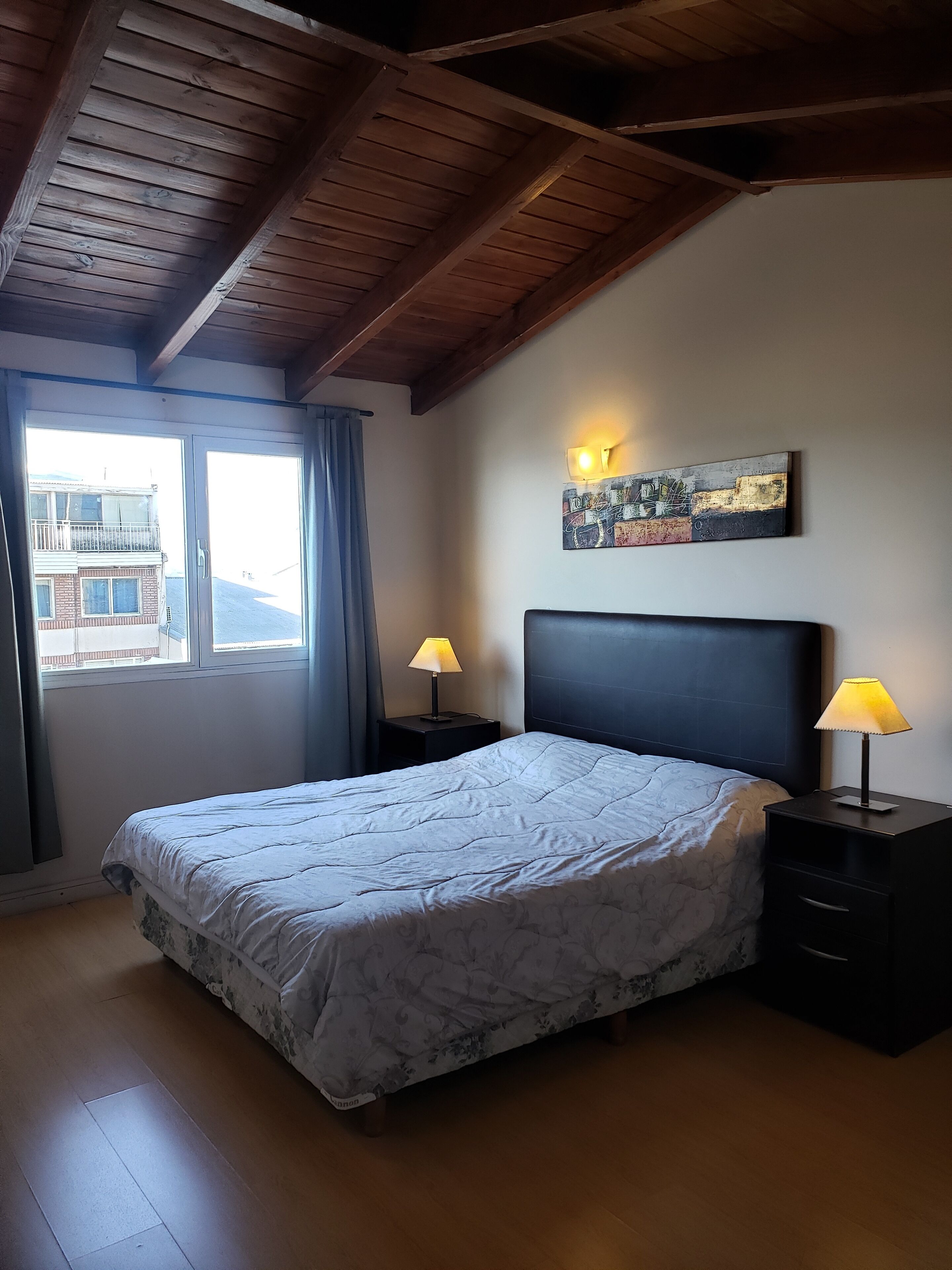 A bedroom with a bed, two bedside tables with lamps, a window with curtains, and a wall-mounted artwork.