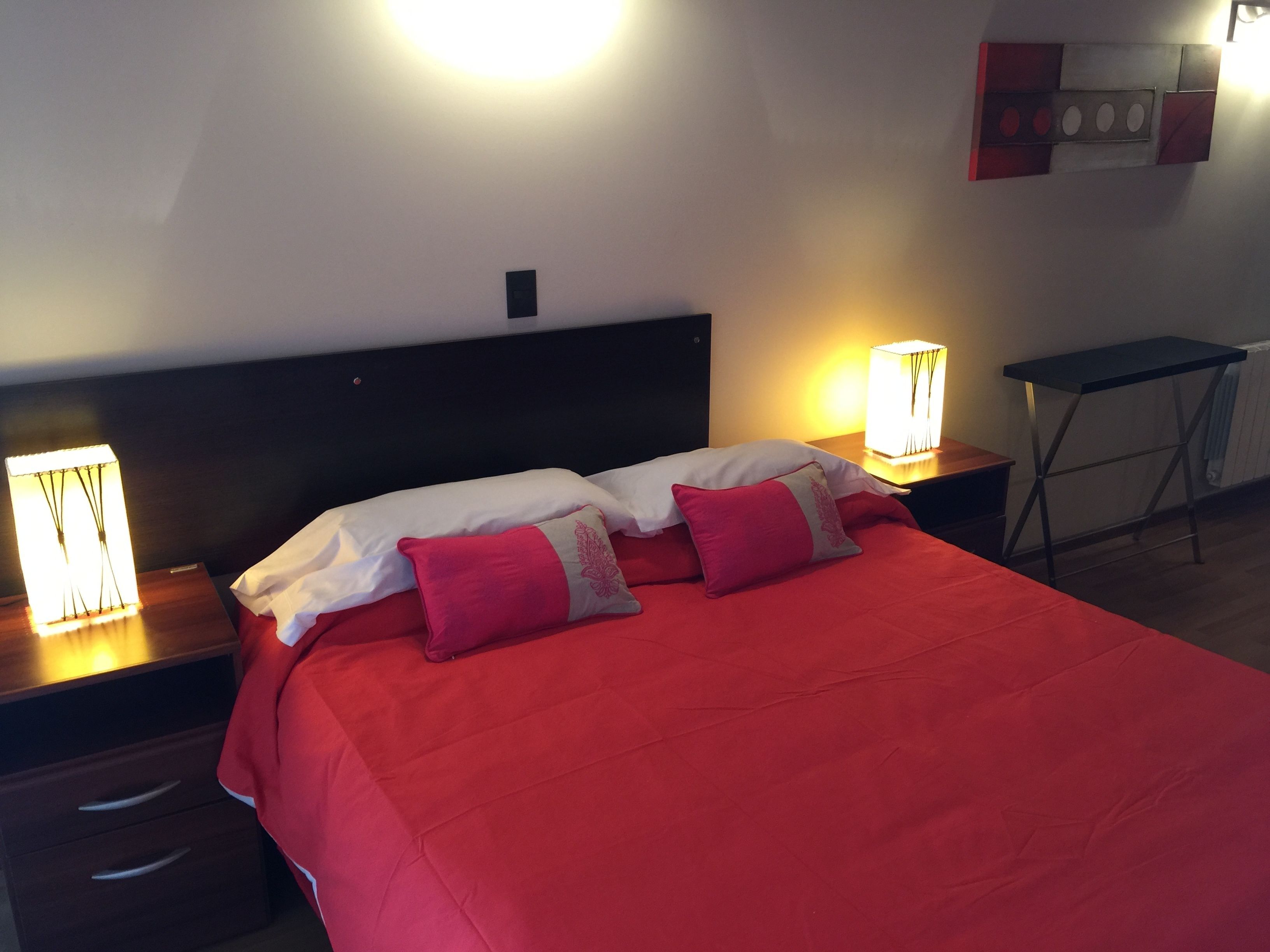 A neatly made bed with red bedding and white pillows, two bedside tables with lamps, a small desk, and a radiator on the wall.