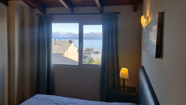 A hotel room with a bed, a nightstand with a lamp, a window with curtains, and a view of mountains and a body of water. - San Martin Suites (San Carlos de Bariloche)