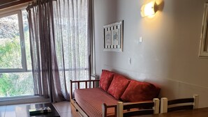 A cozy room with a wooden dining table, a sofa with red cushions, a small black table, and a window with curtains. - San Martin Suites (San Carlos de Bariloche)