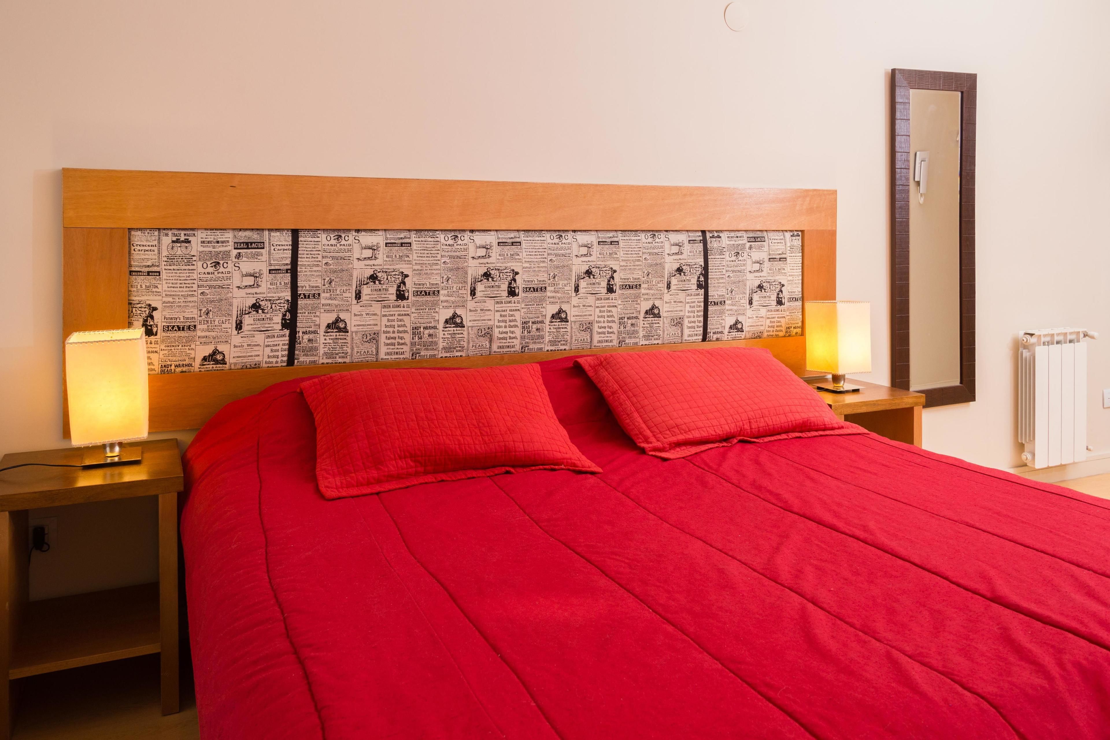 A hotel room with a large bed, red bedding, two bedside lamps, a wooden headboard with newspaper print, a mirror, and a radiator.