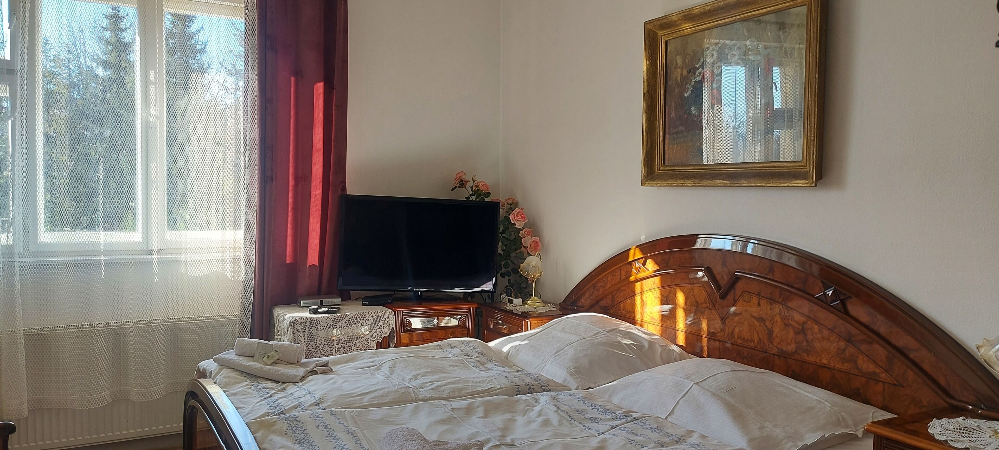 Une chambre à coucher comprenant un lit, une télévision, une table de chevet, un tableau encadré et une fenêtre avec des rideaux.