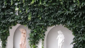 Two white statues in a garden setting with a white wall and green foliage. - Hotel Garni (Ostrava)