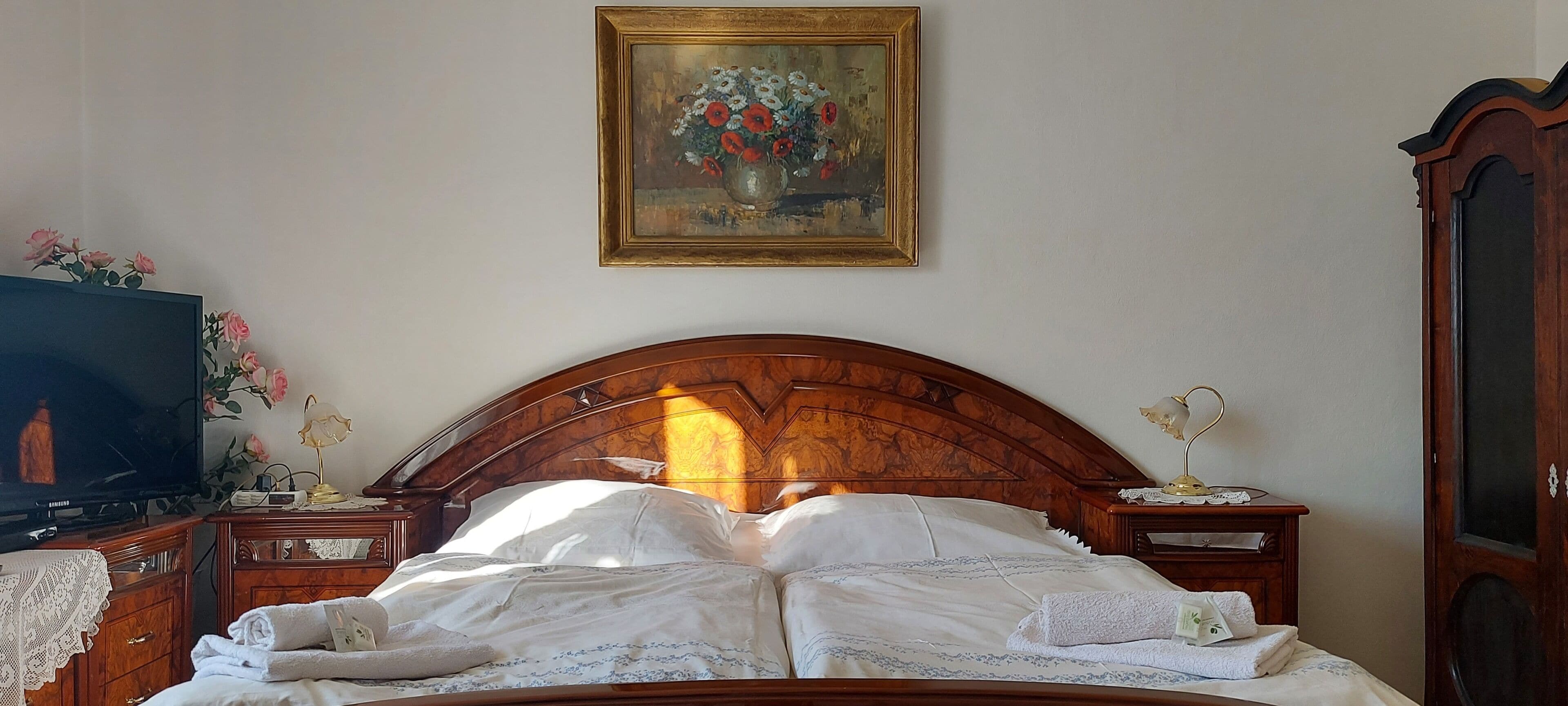 A bedroom with a wooden headboard, a bed with white linens, a painting above the bed, a television on a wooden dresser, and a desk with a lamp.