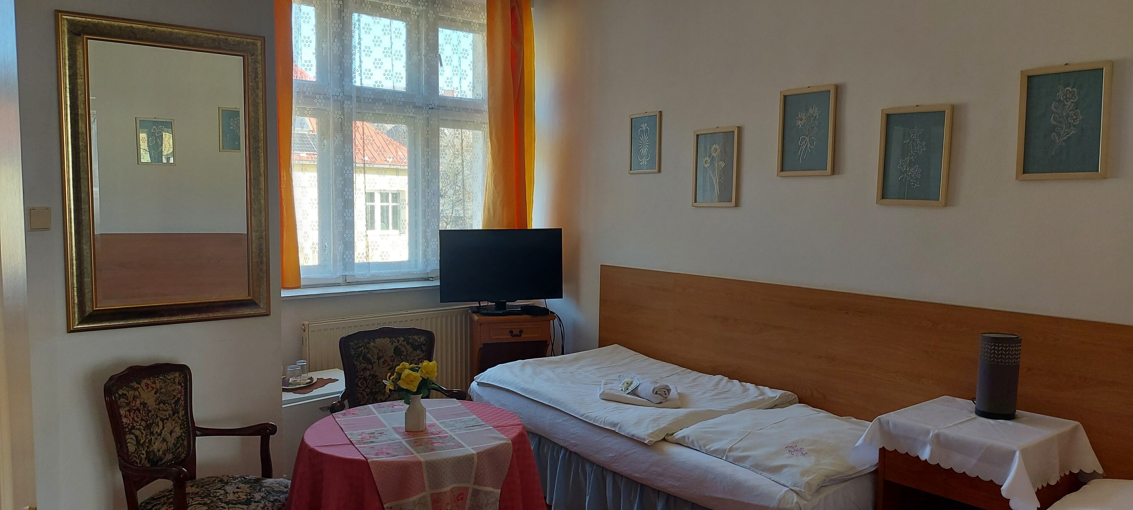 Une chambre d’hôtel avec un lit, une télévision, une chaise, une table ornée d’un vase de fleurs et des tableaux encadrés accrochés au mur.