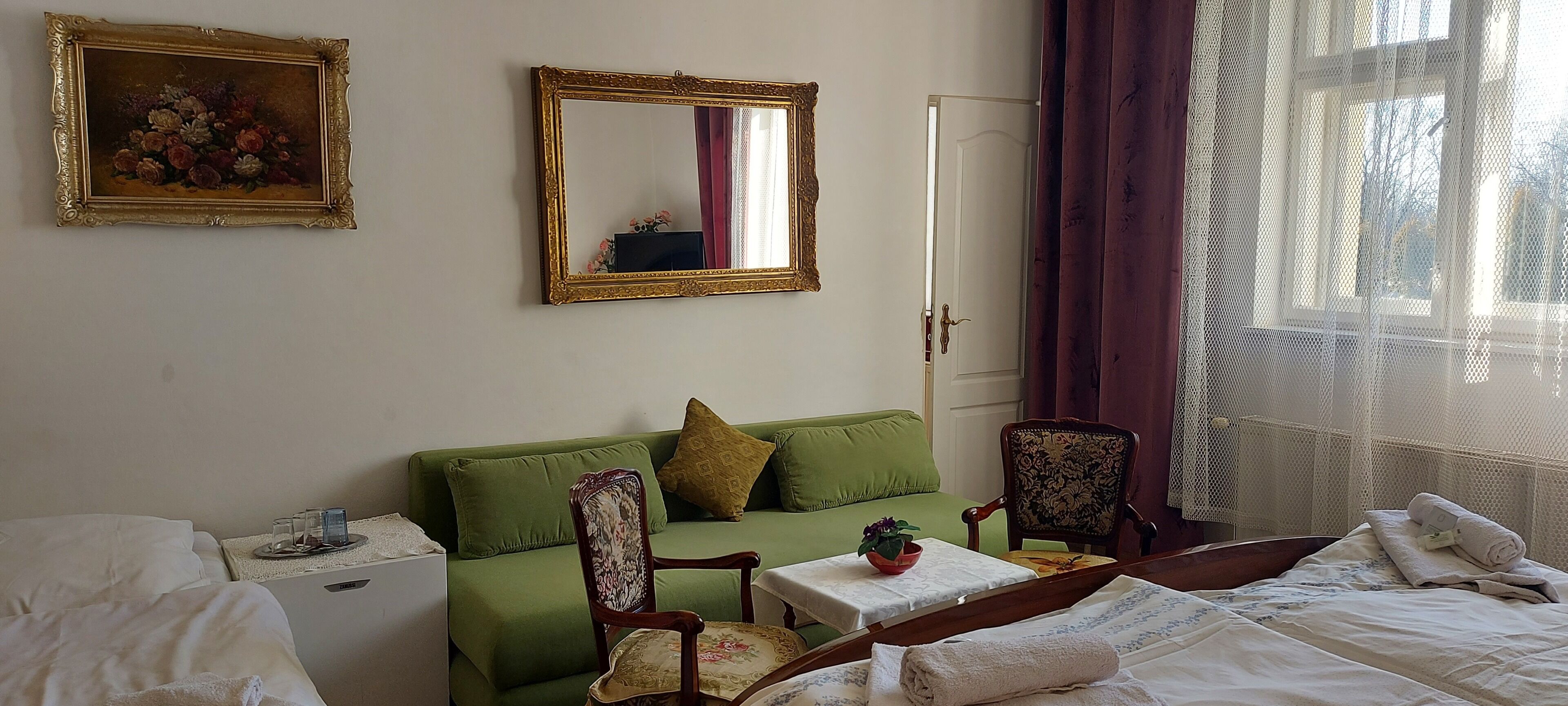 Habitación de hotel con sofá verde, una silla de madera, una mesita con una planta, cama con ropa blanca, un cuadro y un espejo en la pared.