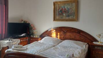 A neatly made bed with a white comforter and pillows, a television on a wooden stand, a painting on the wall, and a nightstand with a lamp.