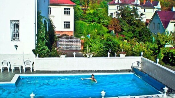 A swimming pool with a person in the water, surrounded by lush greenery and houses. - Hotel Garni (Ostrava)