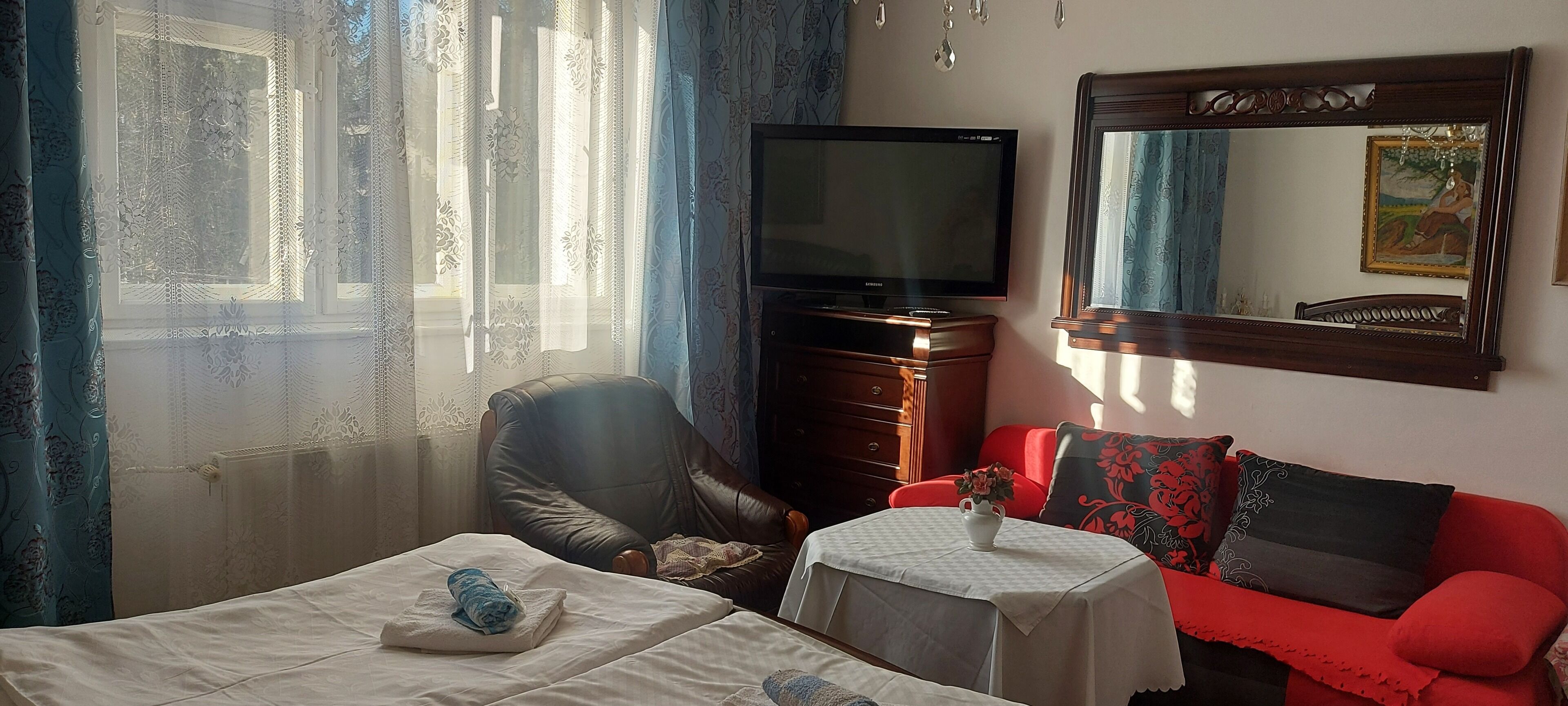A hotel room with a bed, a chair, a small table with a vase, a red sofa, a television, and a large mirror.