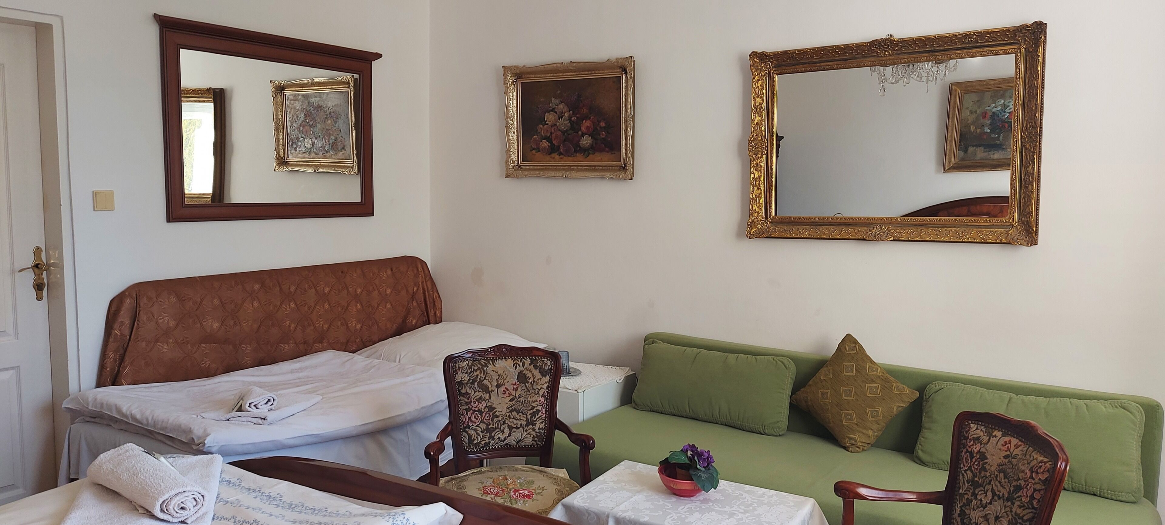 A room with a bed, a green sofa, a wooden chair, a table with a bowl, and three mirrors on the wall.