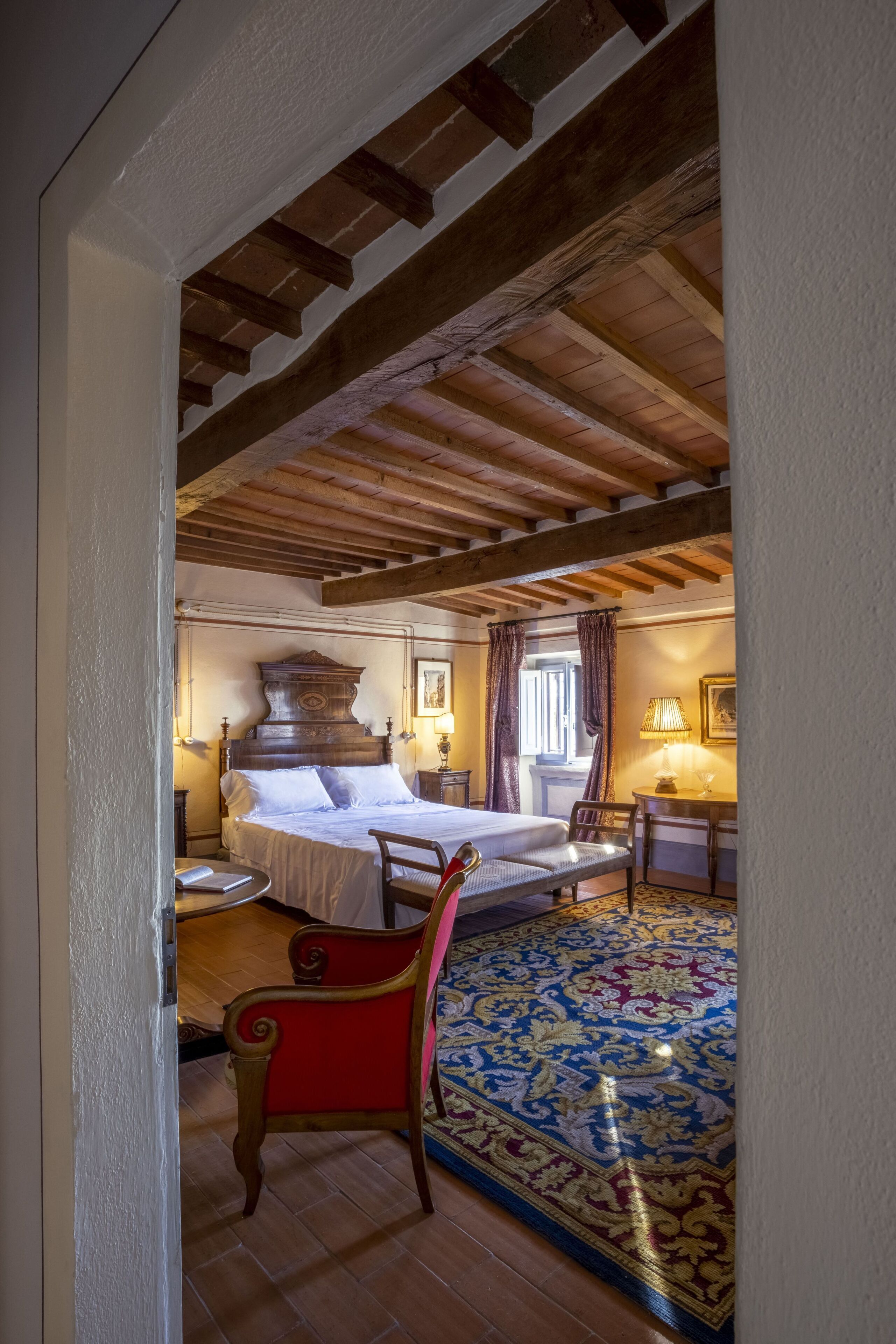A bedroom with a large bed, a wooden ceiling, a red chair, and a patterned rug.