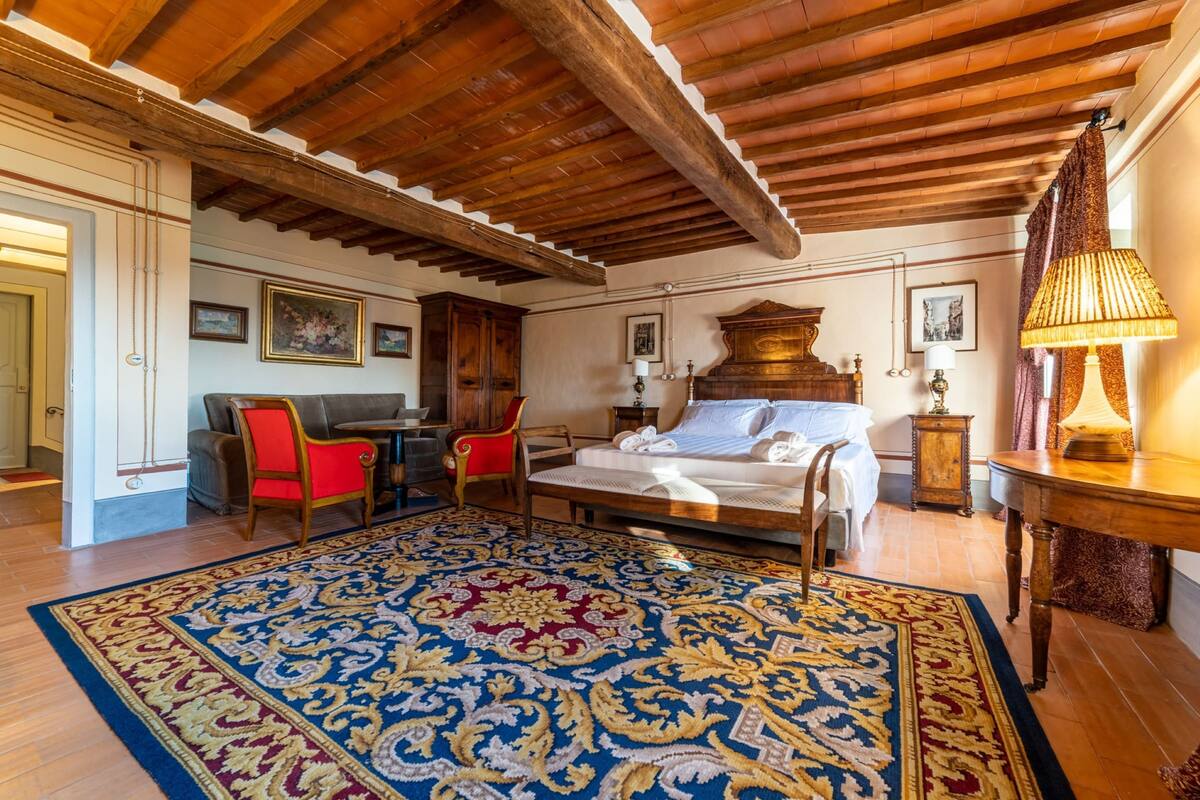 A traditional room with a wooden ceiling, a large bed, a red chair, a desk, and a patterned rug.
