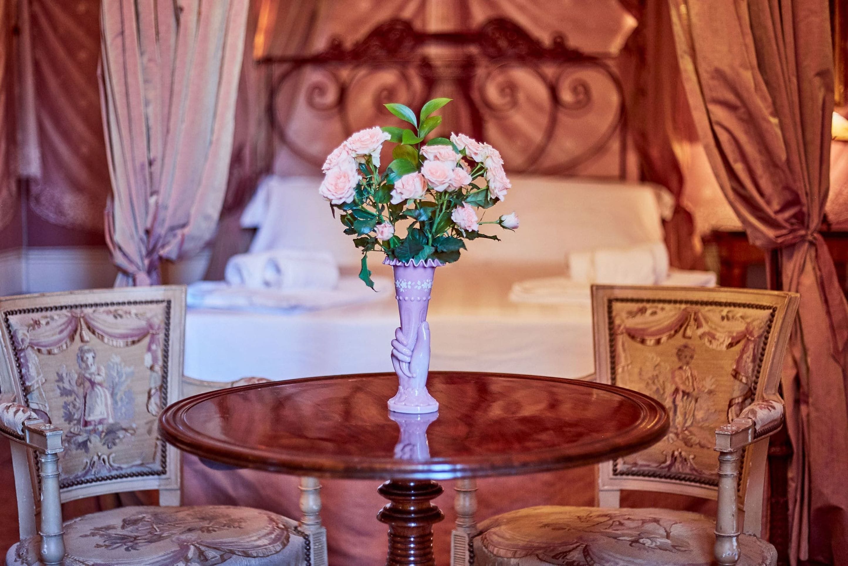 A room with a round table, two ornate chairs, a vase with flowers, and a bed with curtains.