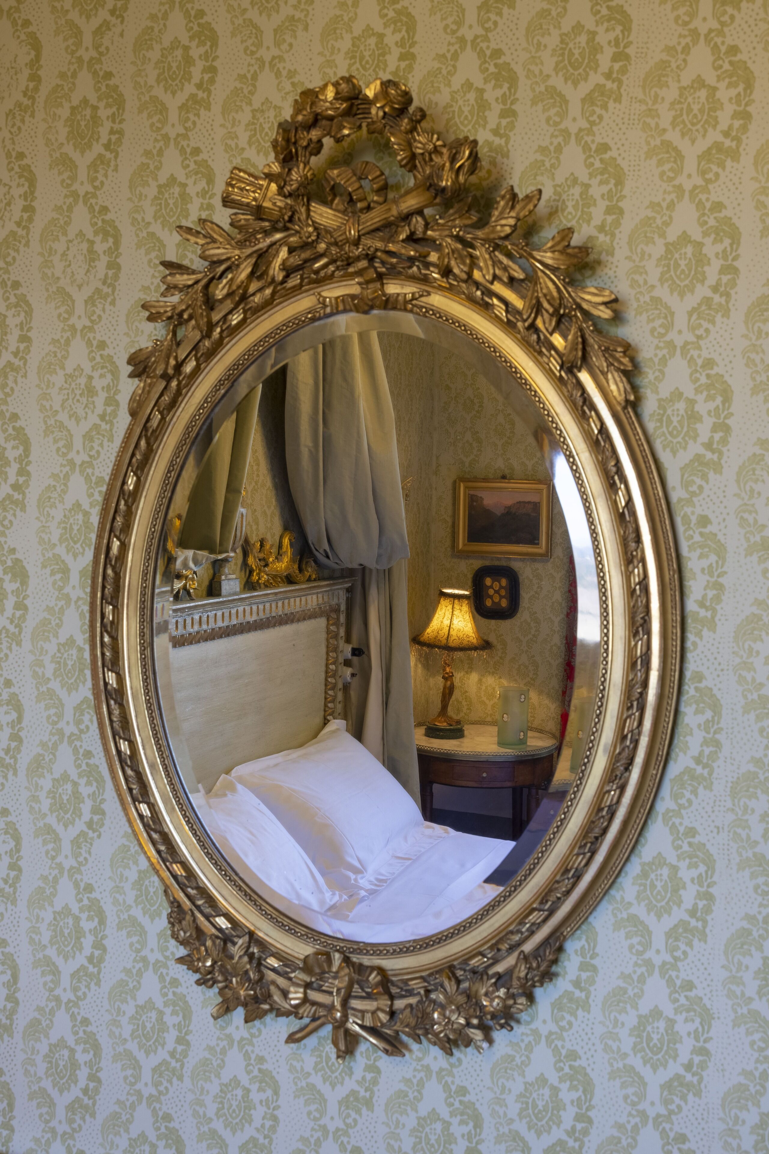 Ein Zimmer mit einem großen, verzierten Spiegel, einem Bett mit weißer Bettwäsche, einem Nachttisch mit Lampe und einem gerahmten Bild an der Wand.