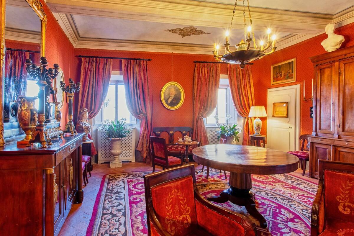 A room with a round wooden table, red upholstered chairs, a chandelier, and a large mirror.