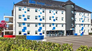 A modern hotel with a white facade and blue accents, featuring a central entrance and a parking lot. - Ibis Budget La Roche sur Yon (Mouilleron-le-Captif)