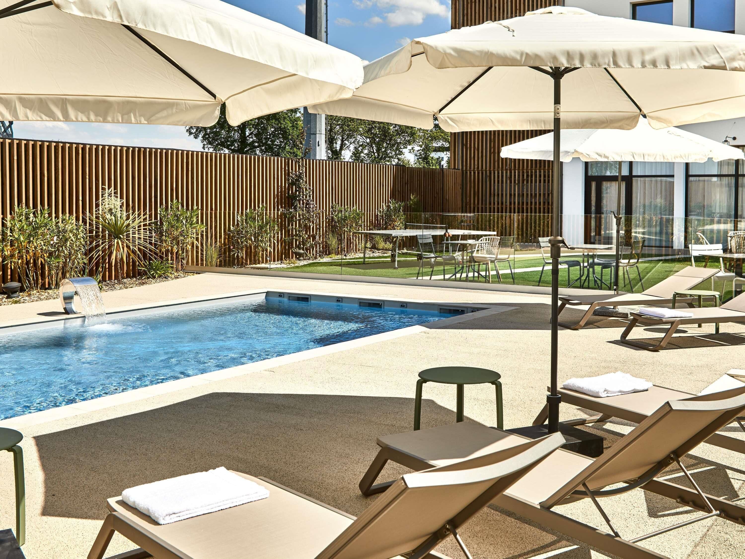 A swimming pool with lounge chairs and umbrellas, surrounded by a wooden fence and greenery.