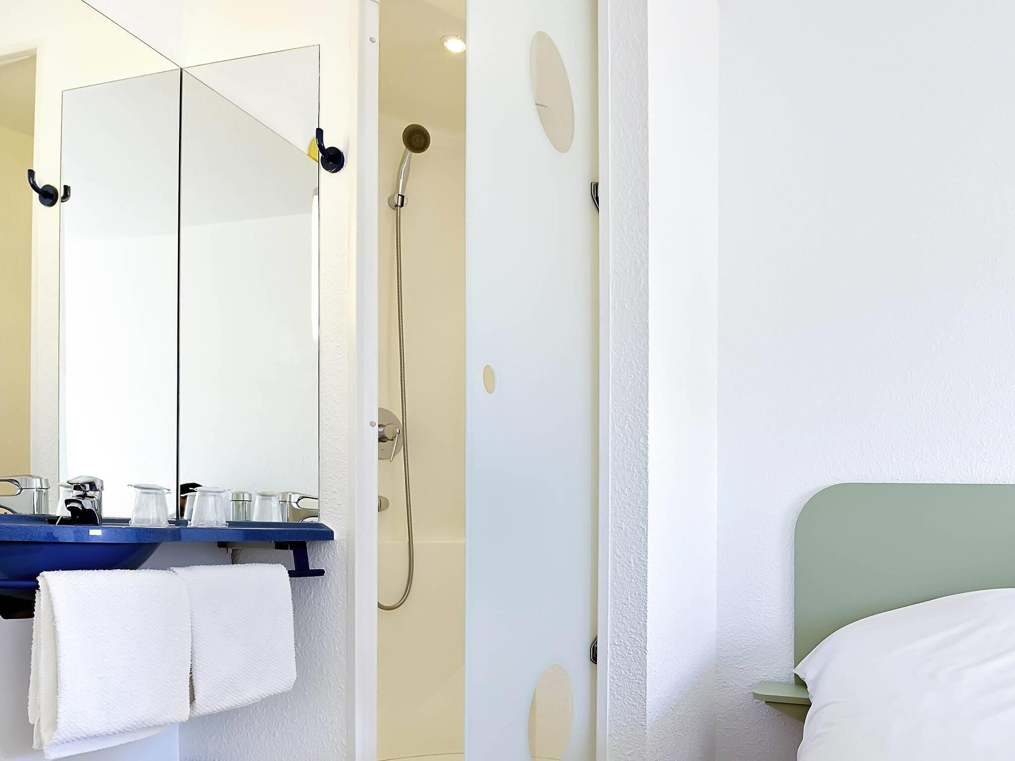 Ein Badezimmer mit Spiegel, Dusche, Waschbecken mit Handtuch und einem Bett mit grünem Kopfteil.