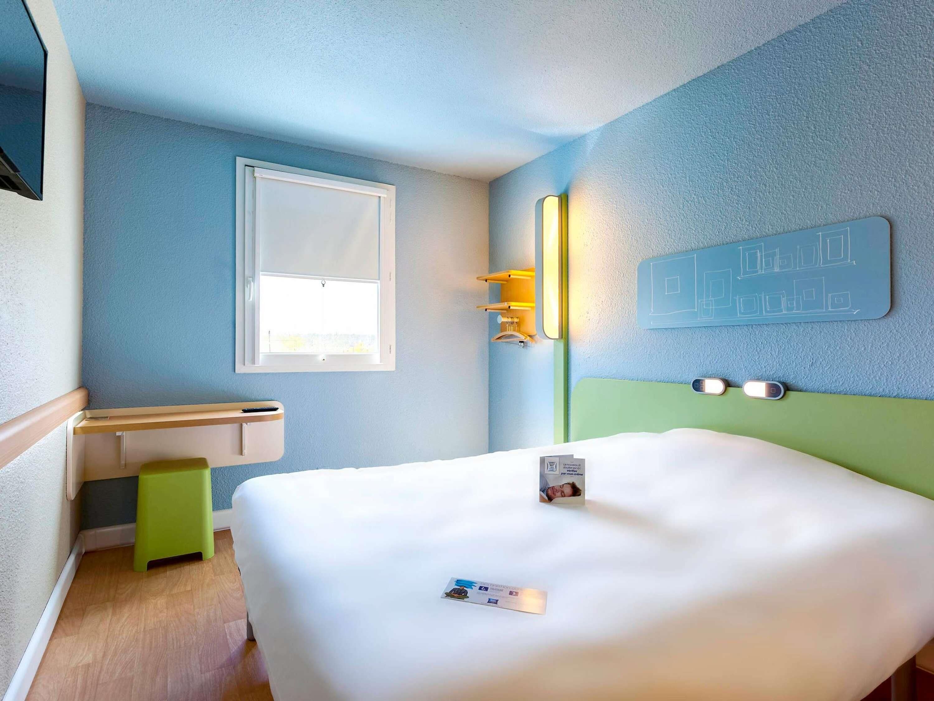 A hotel room with a bed, a window, a wall-mounted light, and a wall decoration.