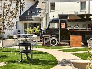 Um food truck antigo com um quadro de menu, mesas e cadeiras ao ar livre, e um edifício com uma placa que diz 'L'Autographe Brasserie' ao fundo.