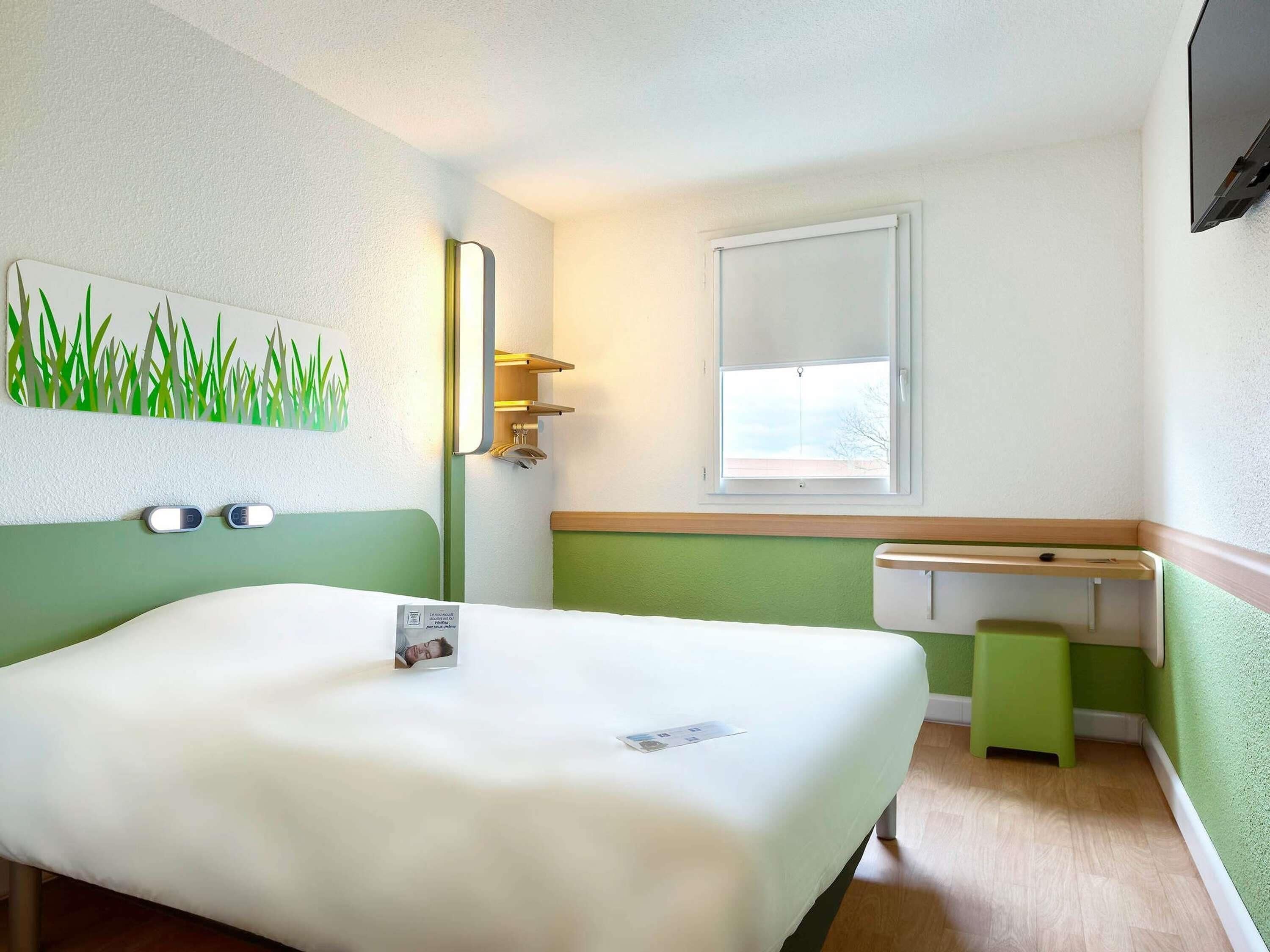 A hotel room with a bed, a small window, a mirror, and a wall-mounted light fixture.