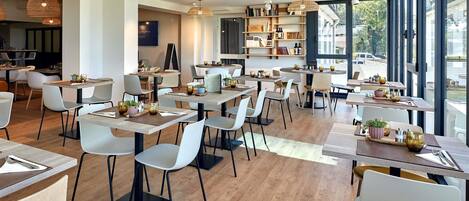 A modern restaurant with wooden tables and chairs, pendant lighting, and large windows.