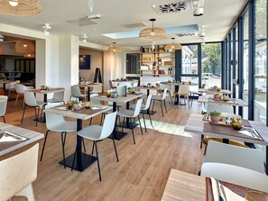 A modern restaurant with wooden tables and chairs, pendant lighting, and large windows.