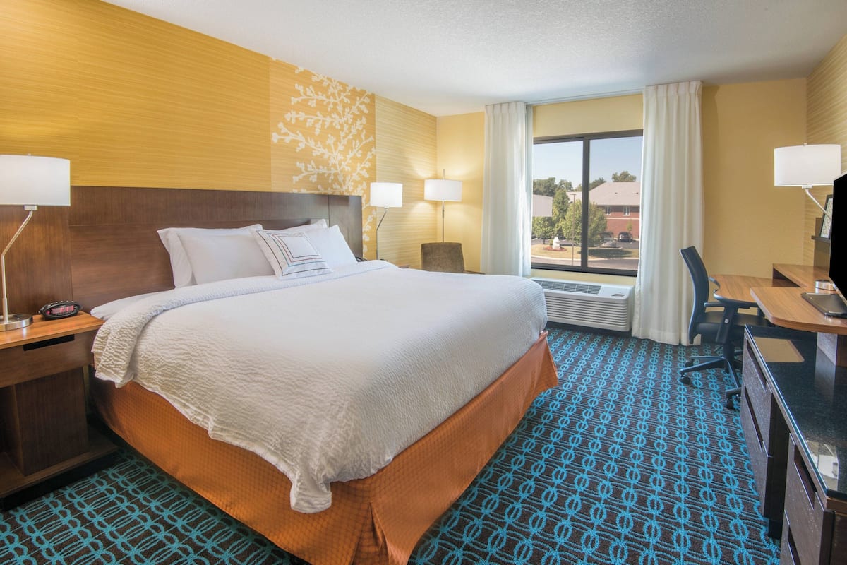 A hotel room with a large bed, two bedside lamps, a desk with a chair, and a view of the outside through a window.